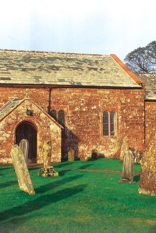 A brick church in a churchyard. There are a number of red stone gravestones in rows diagonally to the front of the church