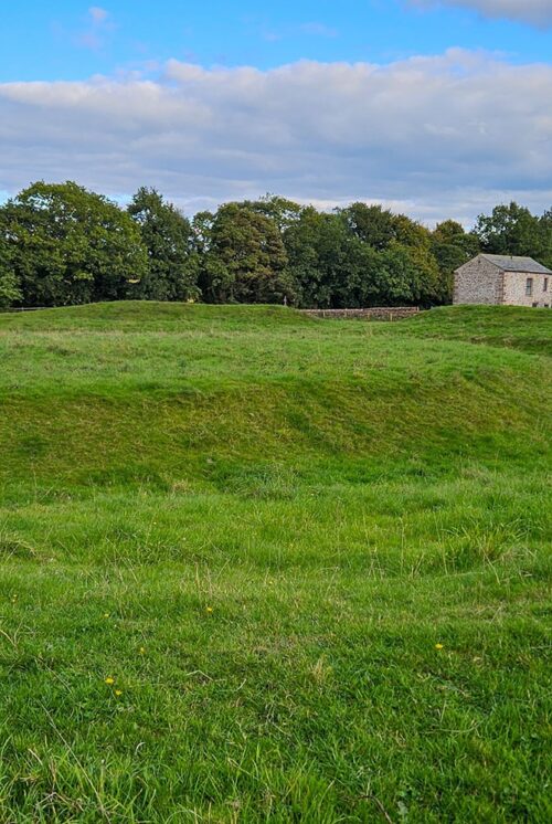 A grassy mound surrounded by a grassy moat in a field. In the background are some stone barns are scattered.