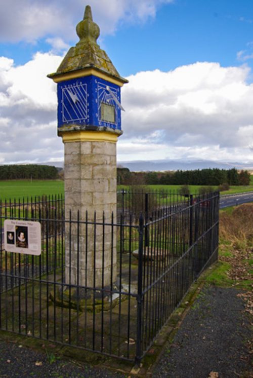 A stone pillar. This is a sundial on one face and on the front face is a square with diagonal lines running through it, both on a blue background.