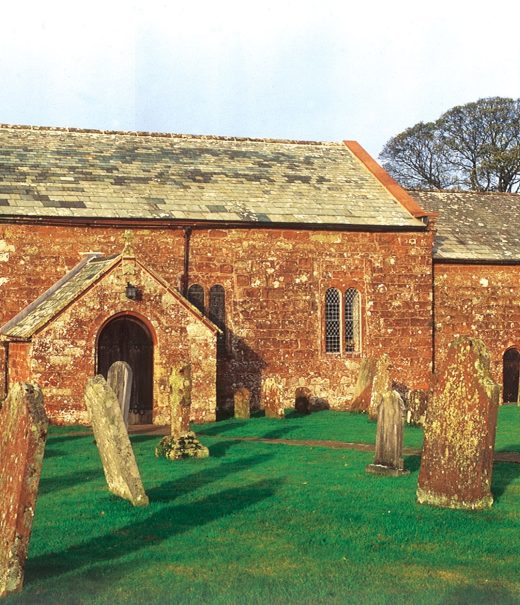 A brick church in a churchyard. There are a number of red stone gravestones in rows diagonally to the front of the church