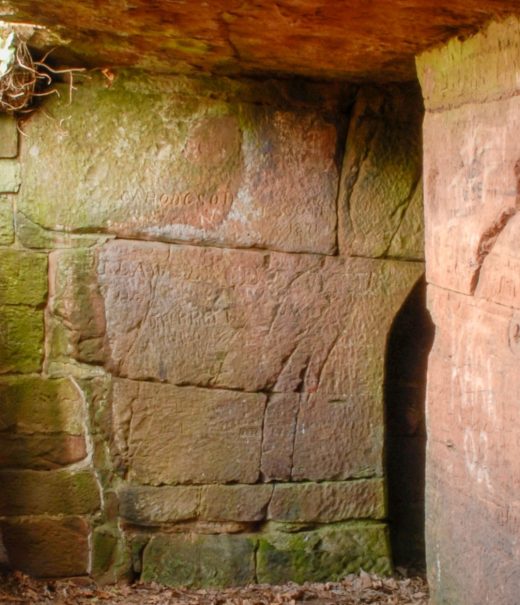 Passageway with a stone overhang that doesn't extend all the way across letting light in. To the right are two entrances to caves that have been carved out of the sandstone.