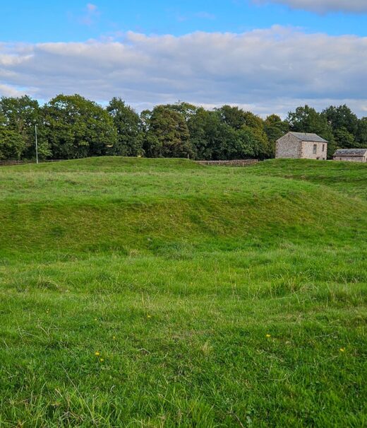 A grassy mound surrounded by a grassy moat in a field. In the background are some stone barns are scattered.