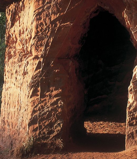 Carved out entrance to red sandstone caves on a cliff overlooking a river.