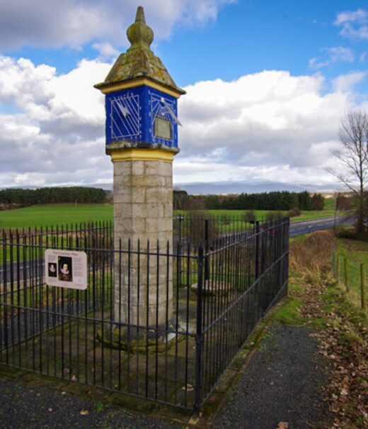 A stone pillar. This is a sundial on one face and on the front face is a square with diagonal lines running through it, both on a blue background.