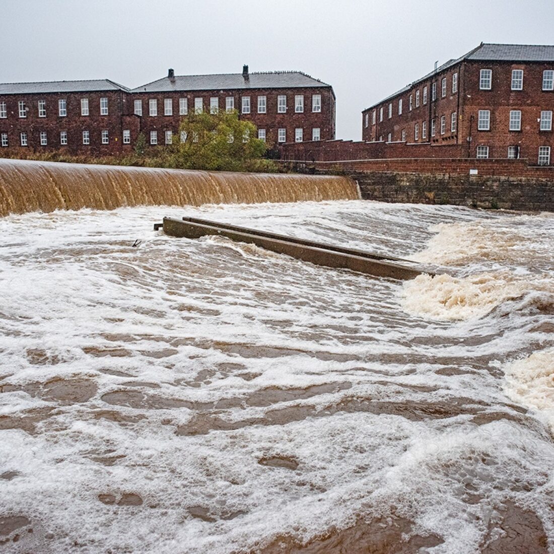 A view across the River Caldew in full spate, cascading over Holme Head weir with buildings behind on far bank at Denton Holme.