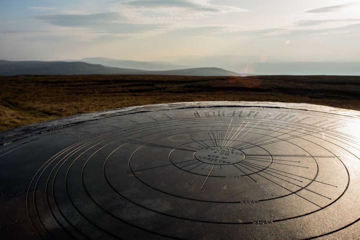Round metal circle on a cairn at the top of the hill. In the distance can see fells. The metal circle has the names and directions of the fells etched onto its surface.