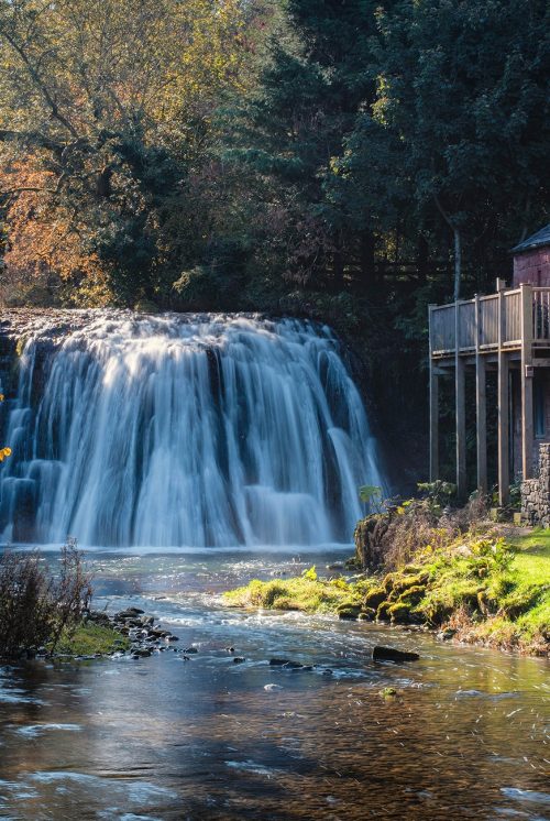 In the background there is a waterfall with water cascading over it in the autumn sunshine. Next to it is an old mill with a wooden waterwheel on the side wall.