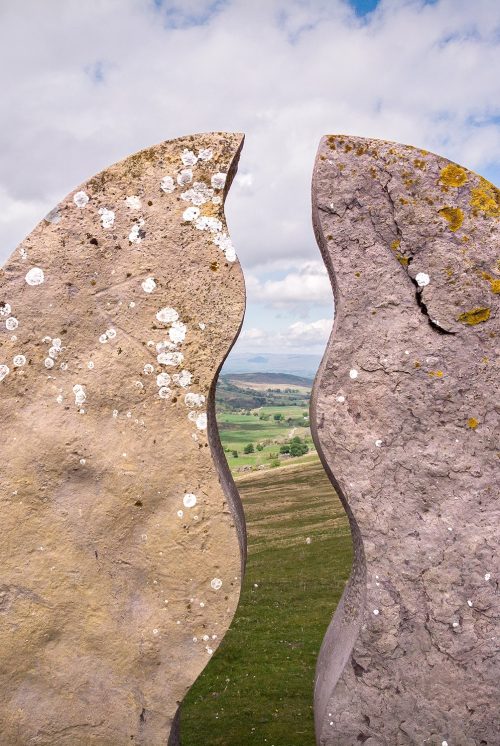 A close-up of a semi-circular stone sculpture that is split in two with wavy lines that look as though a meandering river runs through it. In the background can see the fells and valleys of the Upper Eden in Cumbria.