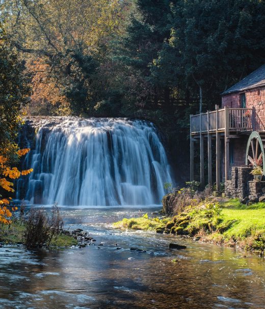 In the background there is a waterfall with water cascading over it in the autumn sunshine. Next to it is an old mill with a wooden waterwheel on the side wall.