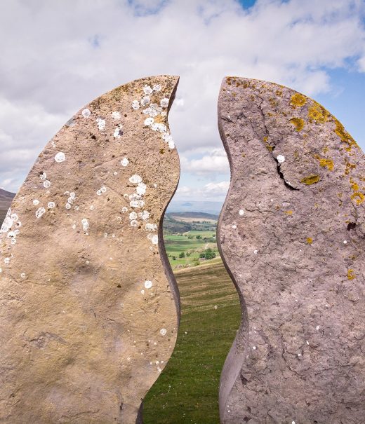 A close-up of a semi-circular stone sculpture that is split in two with wavy lines that look as though a meandering river runs through it. In the background can see the fells and valleys of the Upper Eden in Cumbria.