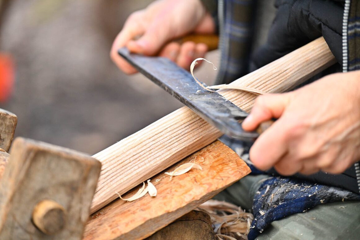 Close up of a tool being used to shave the legs of a wooden bench, tapering them to slot into the bench top.