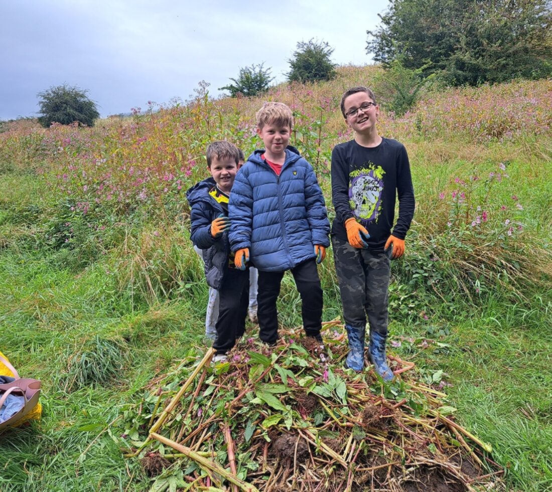 Three young people posing for a photograph standing on a pile of uprooted Himalayan balsam after a day of balsam bashing at Petteril Vale in Carlisle.