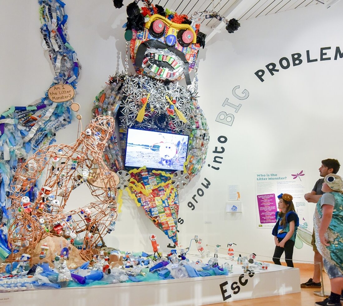 A group of people viewing in an exhibition hall viewing an exhibition piece created from litter depicting a flowing river and a large 'monster' made from discarded items of rubbish.