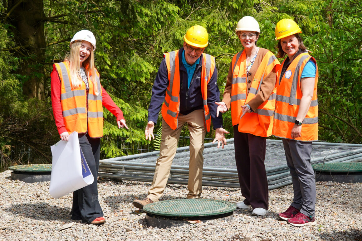 Four people in orange high viz jackets and wearing hard hats are smiling and pointing down at what looks like a manhole cover in the gravel.