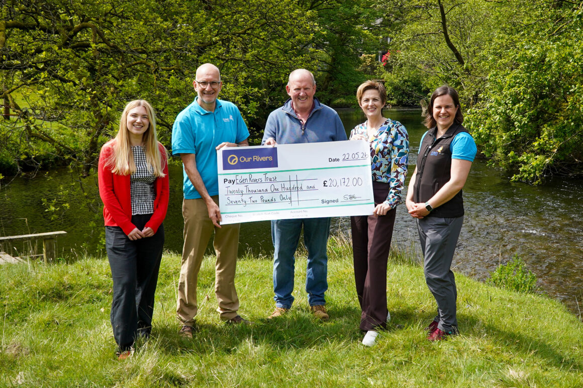 Five people are holding a giant cheque made out to Eden Rivers Trust. They are standing on the riverbank with a small river flowing from left to right behind them