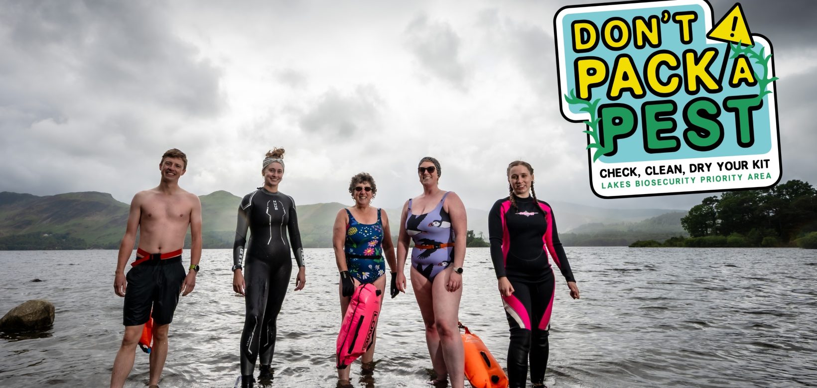Five water users standing in shallow water at the lakes edge. Dressed in shorts, wetsuits and swimming costumes. A graphic in the corner of the image reads: Don't pack a pest.