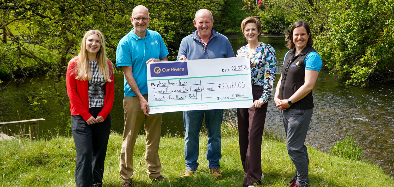 Five people are holding a giant cheque made out to Eden Rivers Trust. They are standing on the riverbank with a small river flowing from left to right behind them