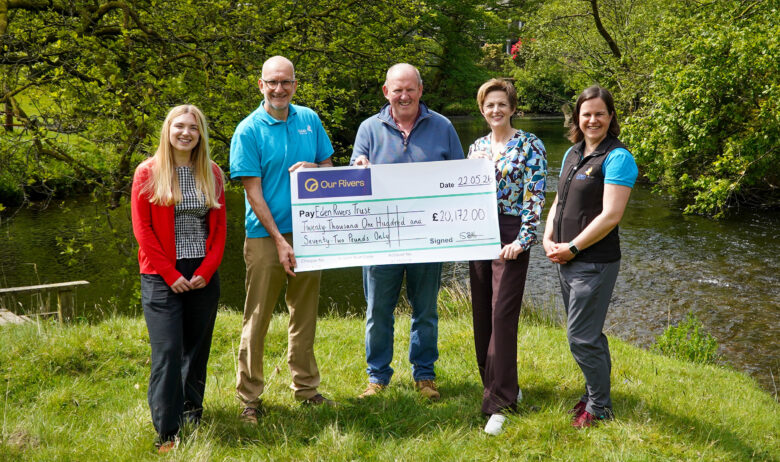 Five people are holding a giant cheque made out to Eden Rivers Trust. They are standing on the riverbank with a small river flowing from left to right behind them