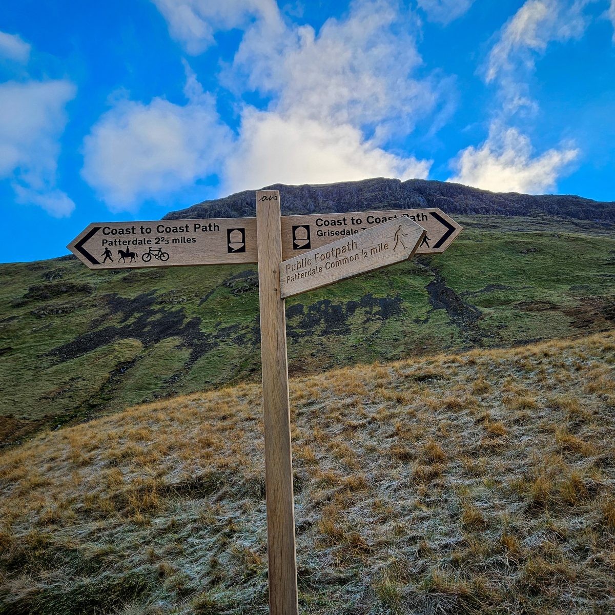 Wooden fingerpost with fellside behind under blue skies. Signpost reads: Coast to Coast path, Patterdale.