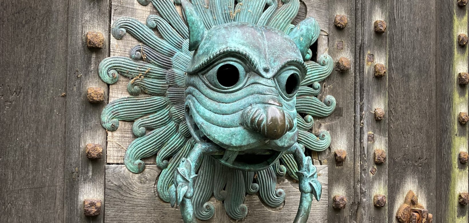 Ornate door knocker in blue oxidised copper in the shape of a lions head. On thick studded wooden door.