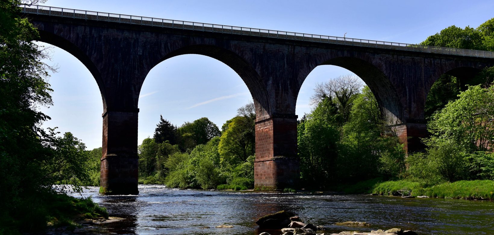 Low level view from wide slow moving river looking up to the arches of a large brick rail viaduct spanning the watercourse.