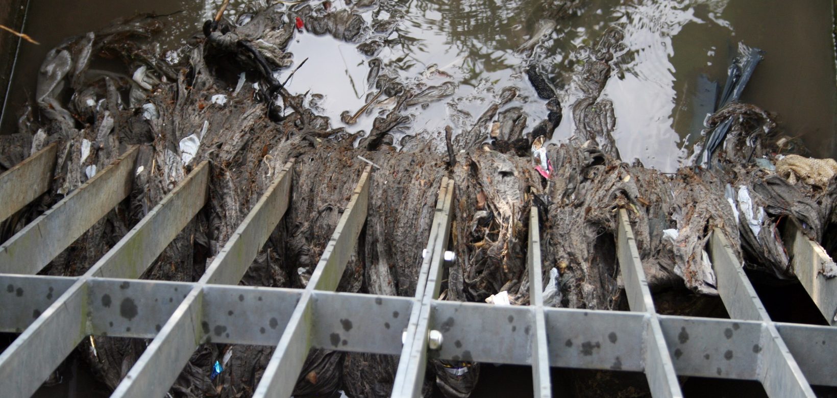 A grill that opens into a river from a sewage pipe. The water is muddy brown and there is discoloured wet wipes and other rubbish stuck in the lower part of the grill.