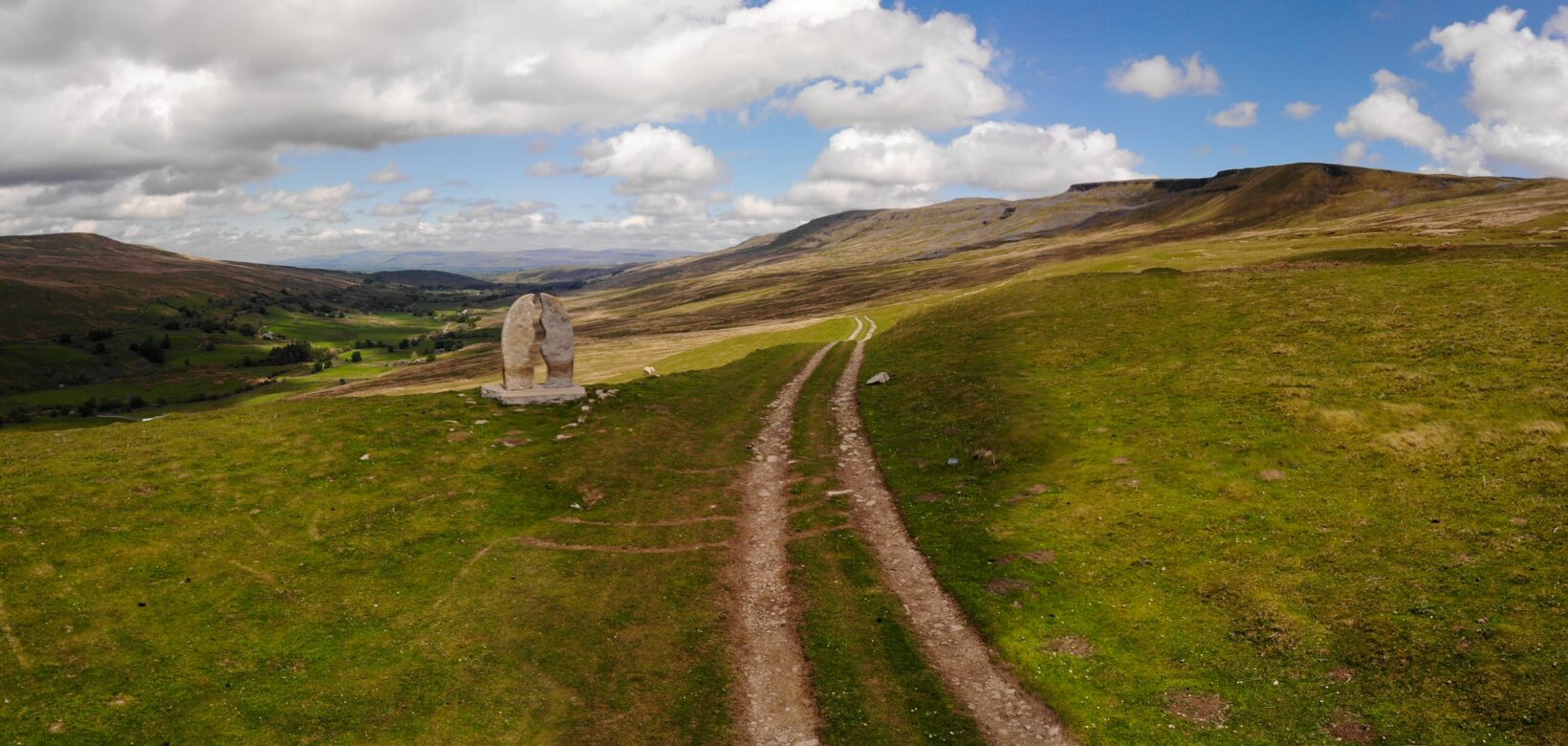 Vehicle track runs across open fell land under blue cloudy skies. A large stone sculpture stands off to the left.