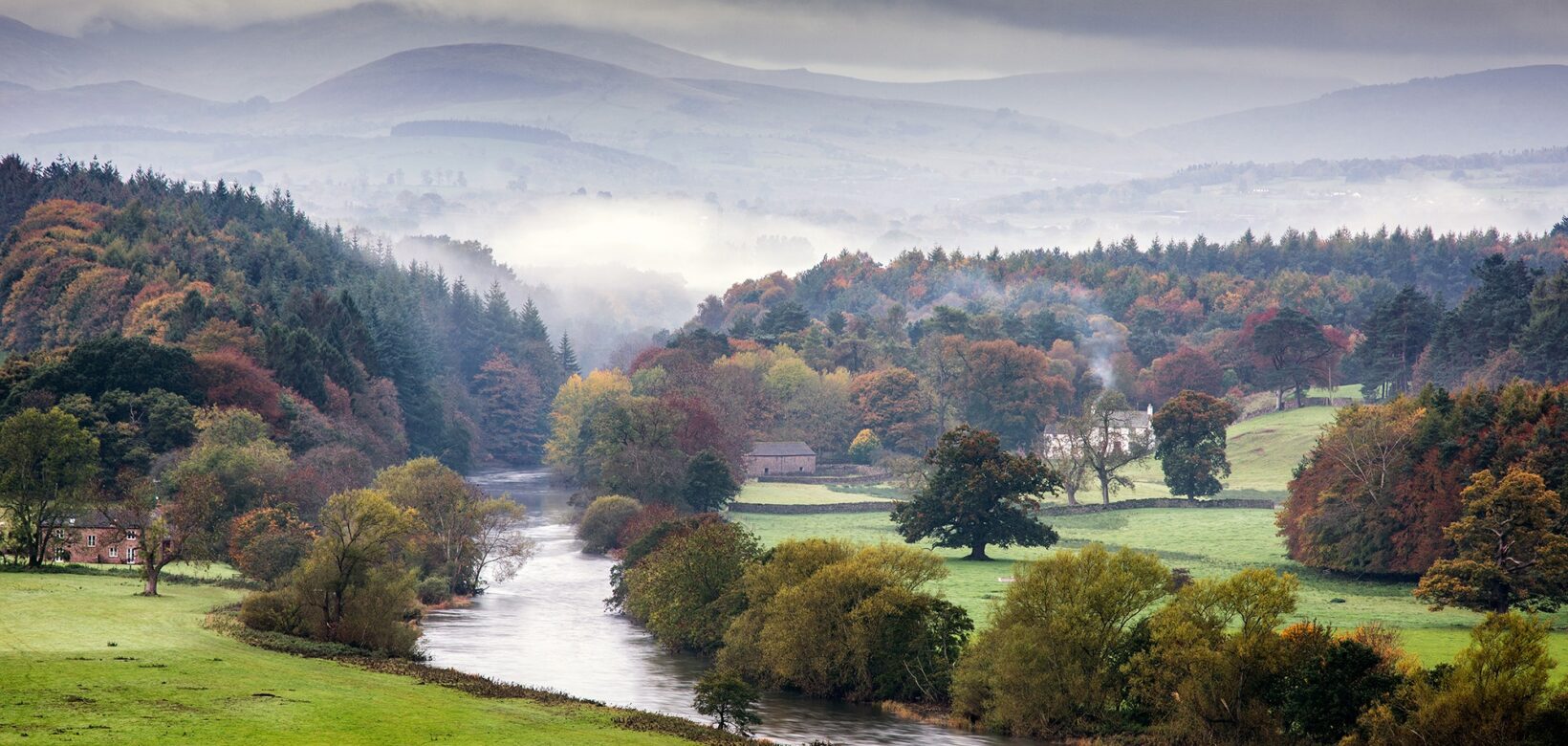 Landscape picture showing a tree-lined river with fields stretching to either side and layers of rolling hills in the background shrouded in mist.