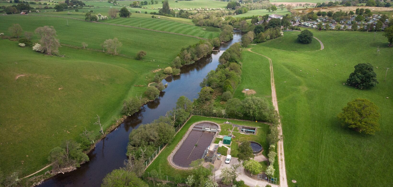 Aerial view of a wide tree lined river running through open grassland dotted with individual mature trees. A small scale water treatment works with stone access pathway sits on the left bank of the watercourse.