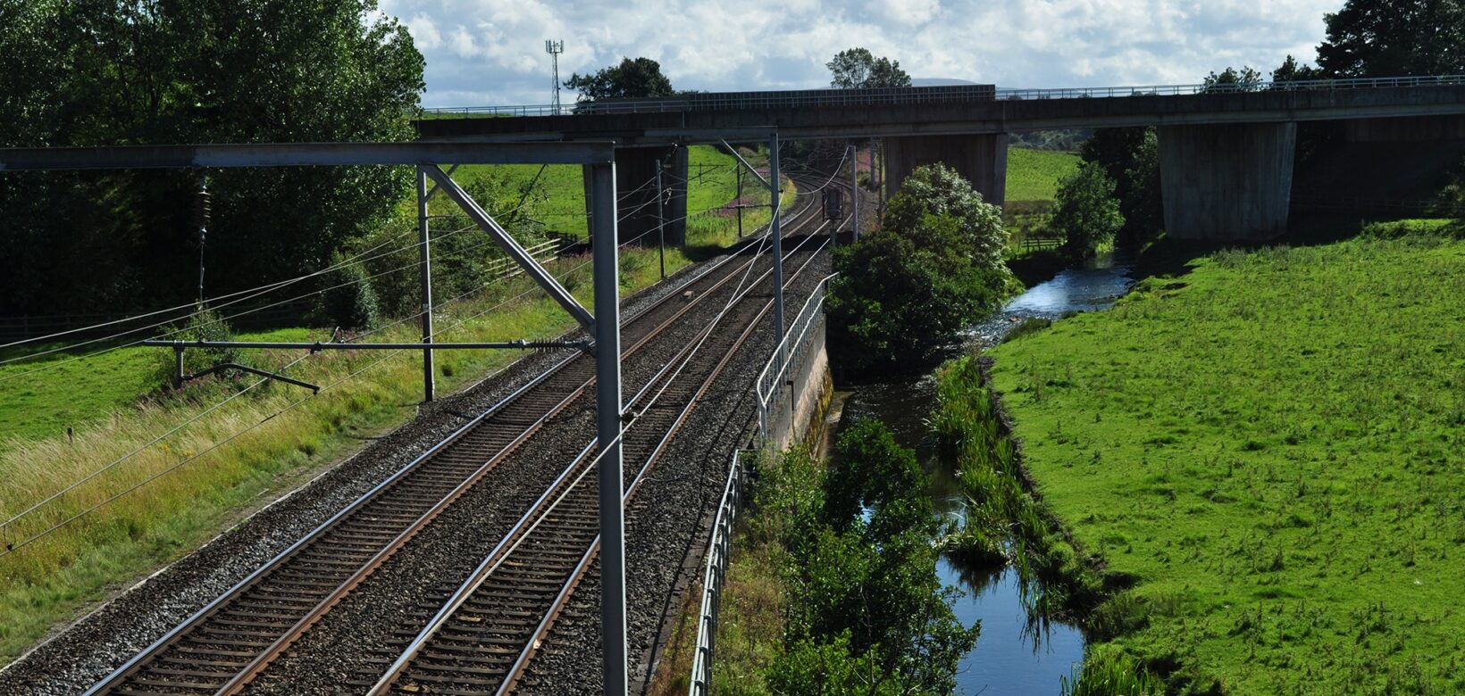 A main railway line runs from front to back of the picture. A river runs along its right-hand side ,and a motorway bridge is at the back of the image, running over both the railway and river.