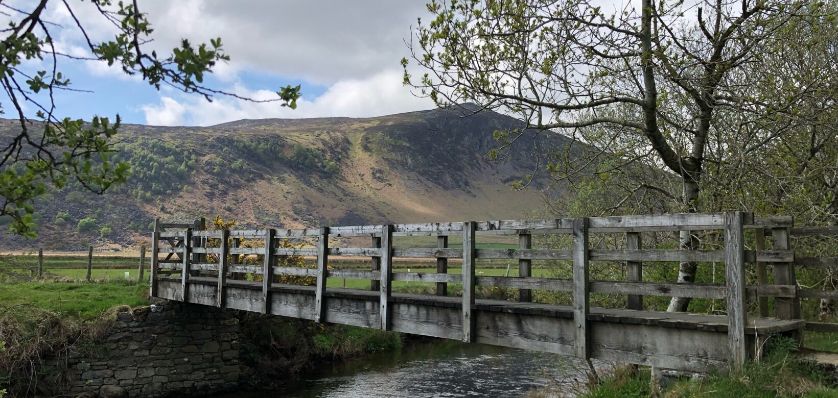 A wooden bridge over a narrow upland part of the River Caldew. In the background a fell can be seen