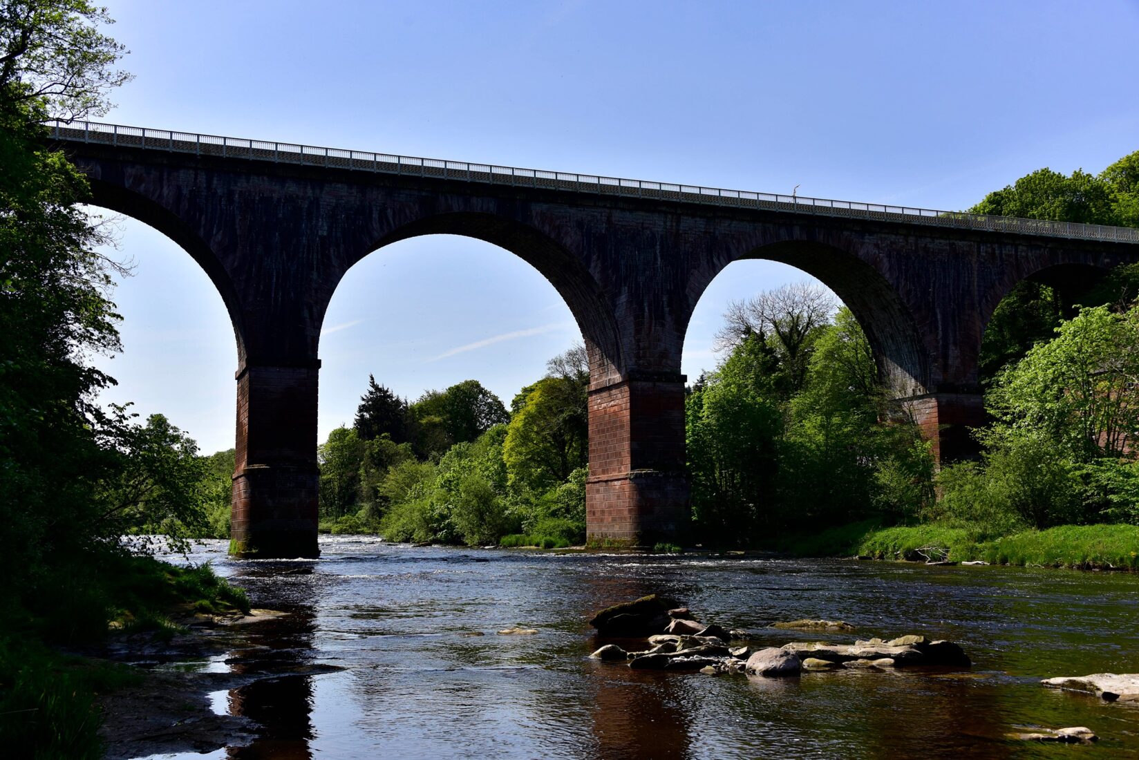 Low level view from wide slow moving river looking up to the arches of a large brick rail viaduct spanning the watercourse.