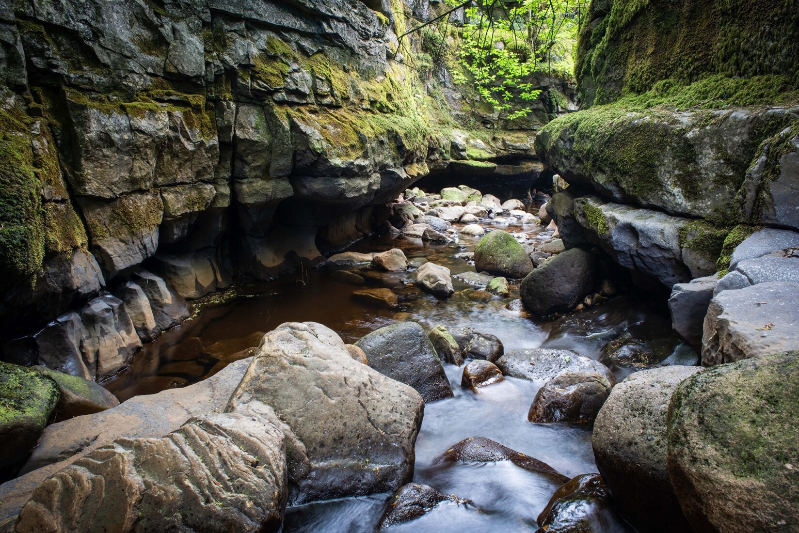 A narrow gorge with deep slow moving water in a boulder strewn base and steep craggy limestone sides towering on either side.
