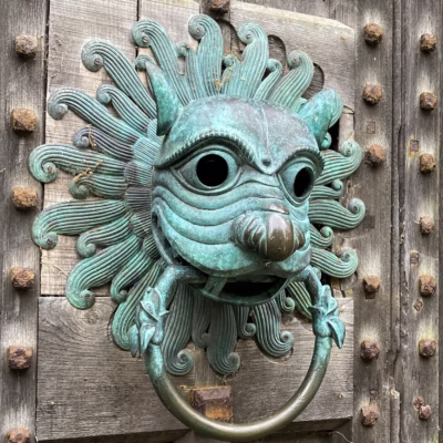 Ornate door knocker in blue oxidised copper in the shape of a lions head. On thick studded wooden door.