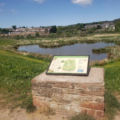 White interpretation board seated atop flat brick platform with Thacka Beck nature reserve lake behind.