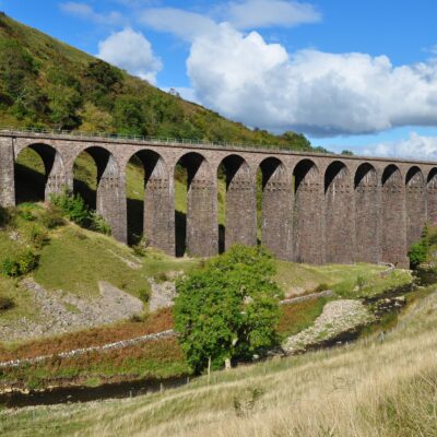 View across grass covered open valley under blue skies to the multiple arches of Smardale viaduct spanning the valley sides beyond.