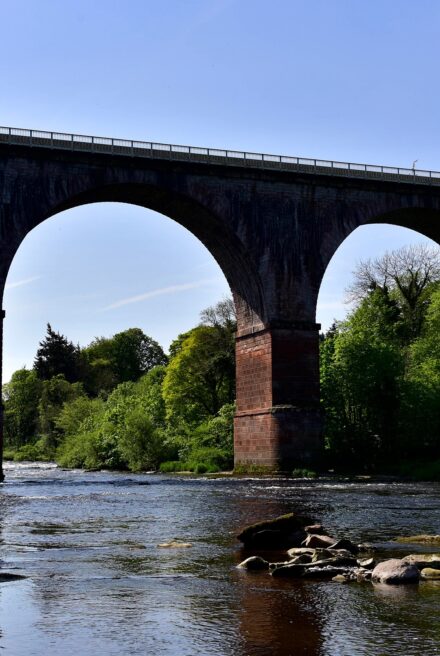 Low level view from wide slow moving river looking up to the arches of a large brick rail viaduct spanning the watercourse.