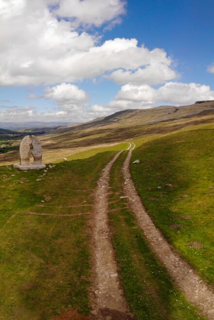Vehicle track runs across open fell land under blue cloudy skies. A large stone sculpture stands off to the left.