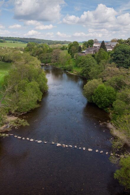 Aerial view of wide river fringed by mixed woodland with grassland beyond. The river is spanned by a line of large boulders forming 'stepping stones' for crossing.
