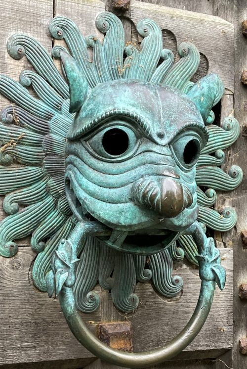 Ornate door knocker in blue oxidised copper in the shape of a lions head. On thick studded wooden door.