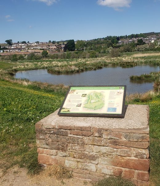 White interpretation board seated atop flat brick platform with Thacka Beck nature reserve lake behind.