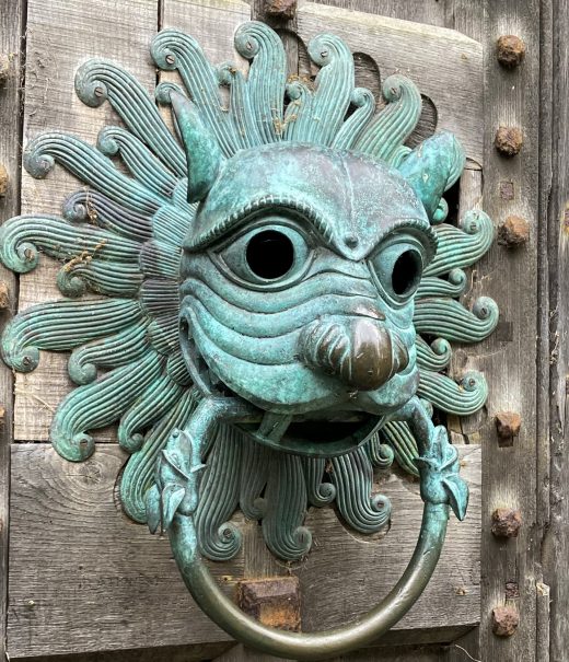 Ornate door knocker in blue oxidised copper in the shape of a lions head. On thick studded wooden door.