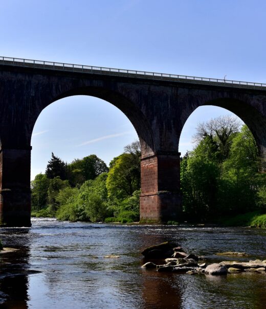 Low level view from wide slow moving river looking up to the arches of a large brick rail viaduct spanning the watercourse.