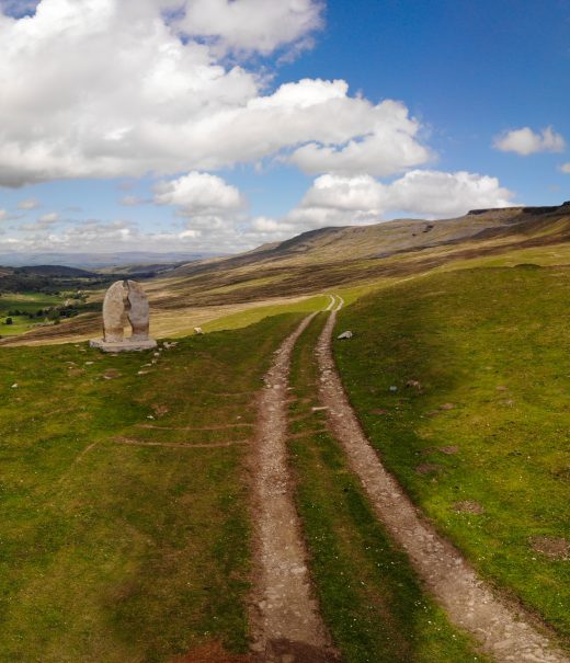 Vehicle track runs across open fell land under blue cloudy skies. A large stone sculpture stands off to the left.