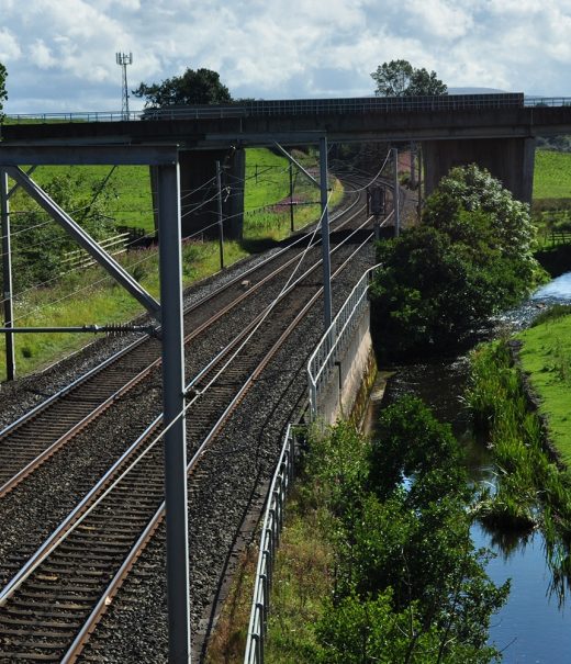 A main railway line runs from front to back of the picture. A river runs along its right-hand side ,and a motorway bridge is at the back of the image, running over both the railway and river.