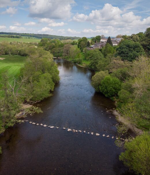 Aerial view of wide river fringed by mixed woodland with grassland beyond. The river is spanned by a line of large boulders forming 'stepping stones' for crossing.