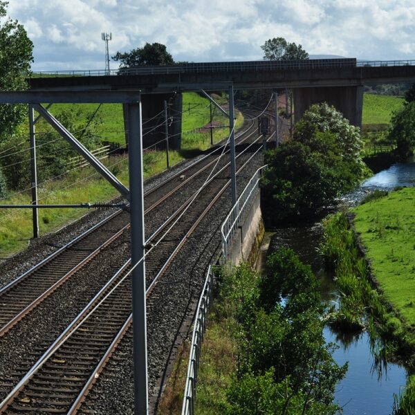 A main railway line runs from front to back of the picture. A river runs along its right-hand side ,and a motorway bridge is at the back of the image, running over both the railway and river.