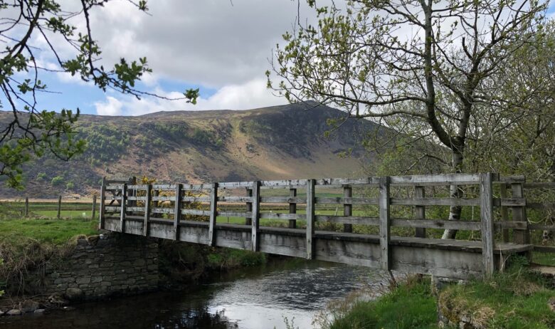 A wooden bridge over a narrow upland part of the River Caldew. In the background a fell can be seen