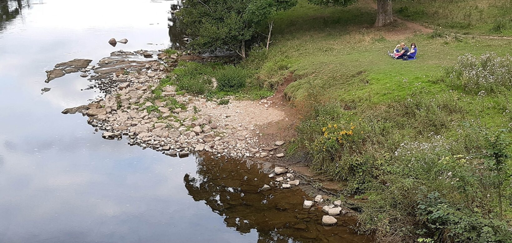 An aerial view of a wide a slow moving section of the river Eden. A stone a boulder shelf encroaches into the river on the inner bank of a slight bend. To the right a small grass bank with two people having a picnic.