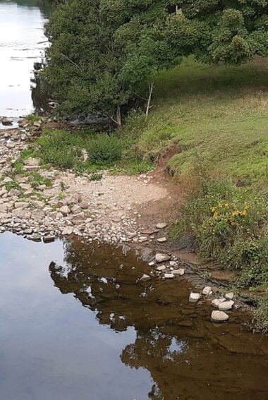 An aerial view of a wide a slow moving section of the river Eden. A stone a boulder shelf encroaches into the river on the inner bank of a slight bend. To the right a small grass bank with two people having a picnic.